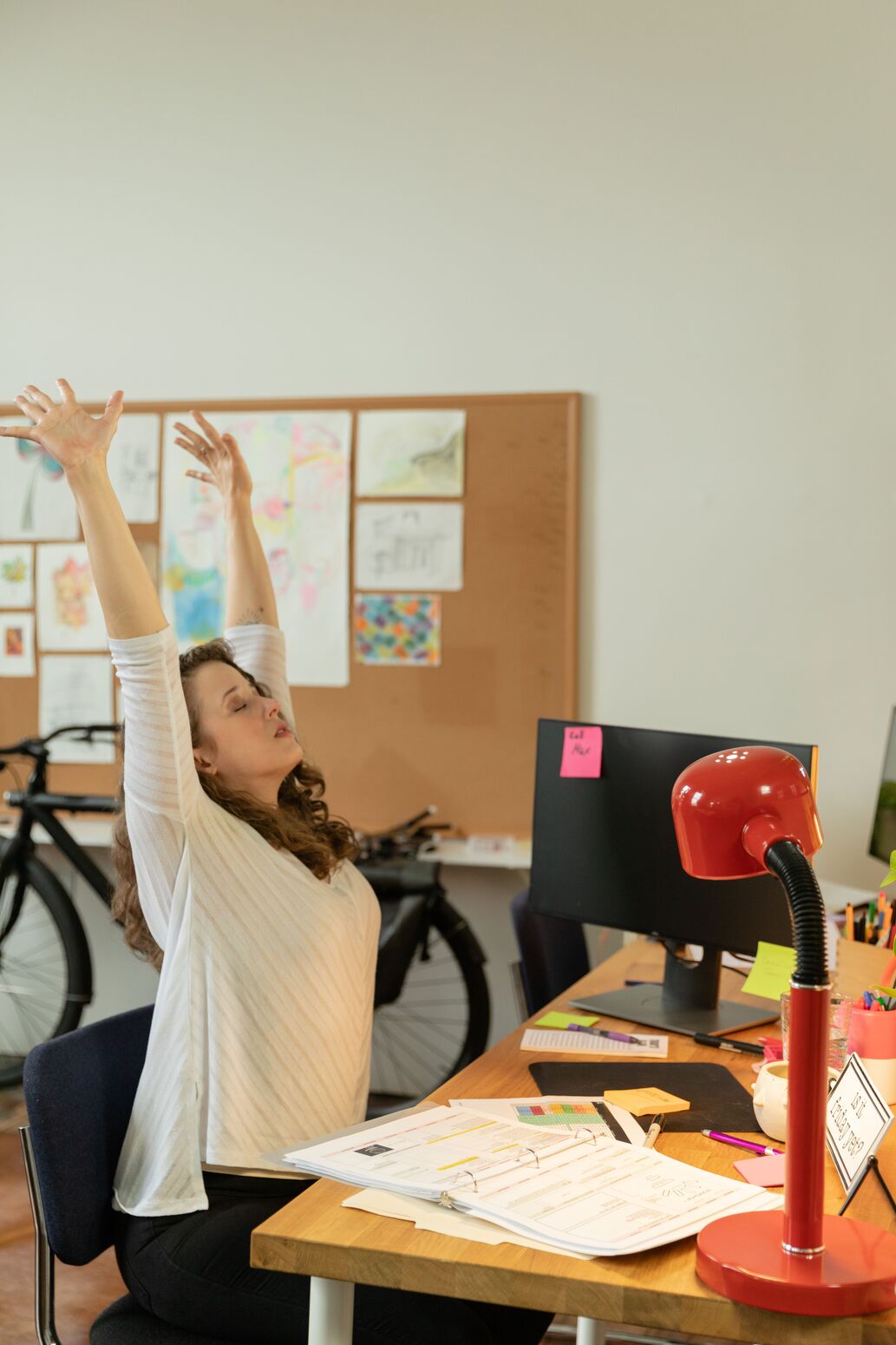 Woman at a desk with a red lamp stretching both arms overhead during a work break