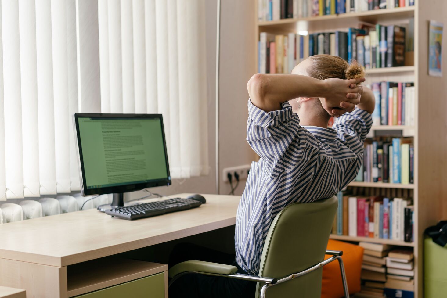 Woman in a striped shirt on an office chair with hands behind her head, looking at a monitor